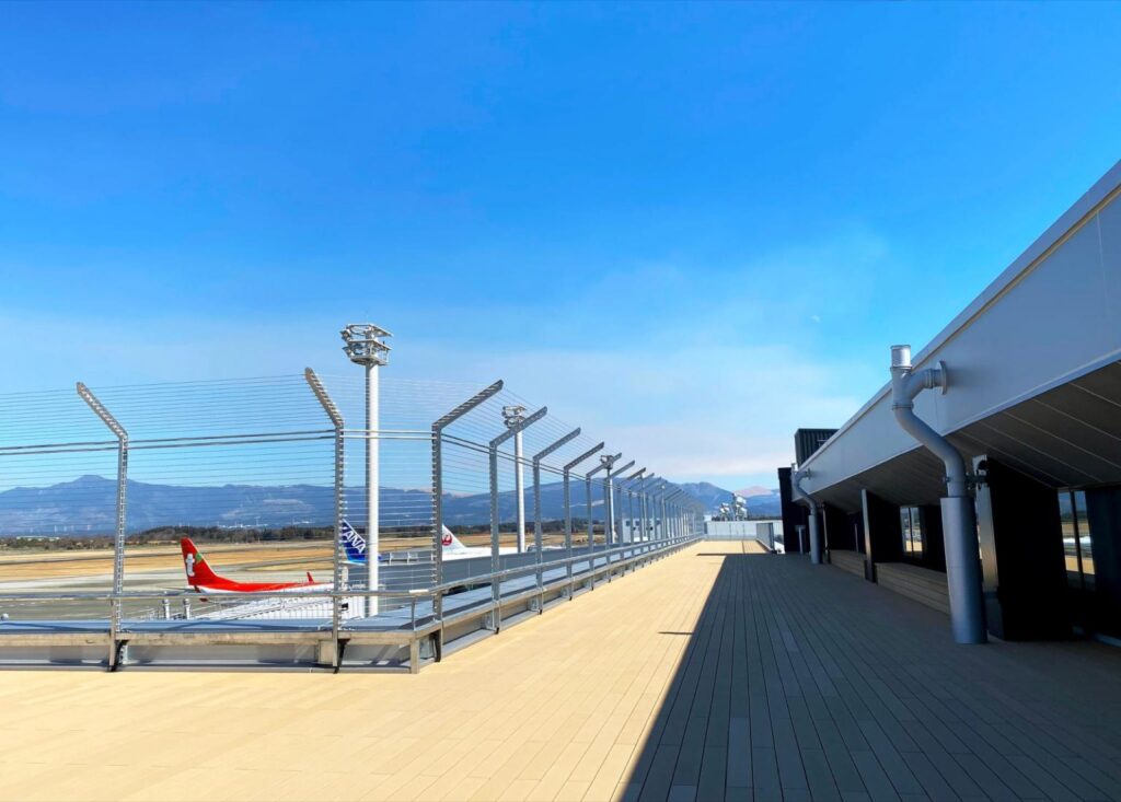 An outdoor deck at the terminal of Aso Kumamoto Airport, with aircraft and mountains beyond the fence